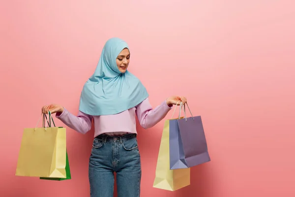 Young muslim woman in hijab and jeans holding multicolored shopping bags on pink background — Stock Photo