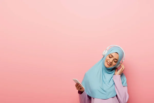 Happy muslim woman holding cellphone while listening music in headphones isolated on pink — Stock Photo