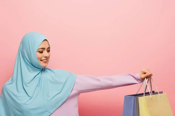Cheerful arabian woman in blue hijab showing multicolored shopping bags on pink background — Stock Photo