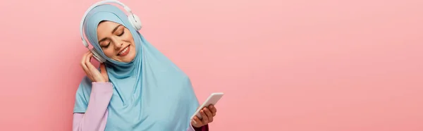 Mulher árabe positivo em azul hijab ouvir música em fones de ouvido sem fio isolados em rosa, banner — Fotografia de Stock