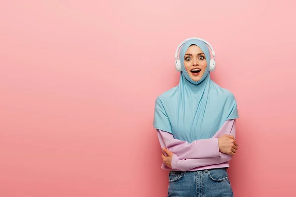 Donna araba stupita in hijab blu e cuffie guardando la fotocamera su sfondo rosa — Foto stock