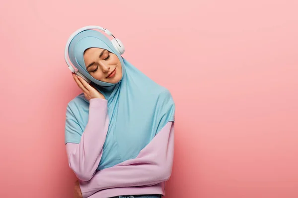 Mujer musulmana complacida escuchando música en auriculares con los ojos cerrados sobre fondo rosa - foto de stock