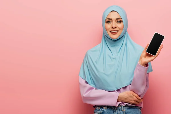 Mujer árabe bonita sonriendo a la cámara mientras muestra el teléfono móvil con pantalla en blanco sobre fondo rosa - foto de stock