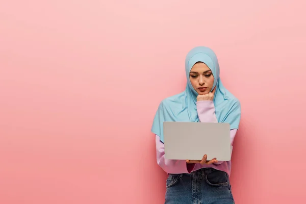 Вдумчивая мусульманка смотрит на ноутбук на розовом фоне — стоковое фото