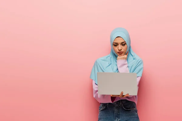 Nachdenkliche muslimische Frau schaut auf Laptop auf rosa Hintergrund — Stockfoto