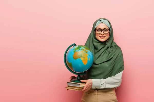 Insegnante musulmano sorridente con libri di testo e globo guardando la fotocamera su sfondo rosa — Foto stock