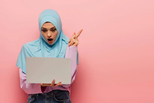 Surprised muslim woman pointing with finger while looking at laptop on pink background — Stock Photo