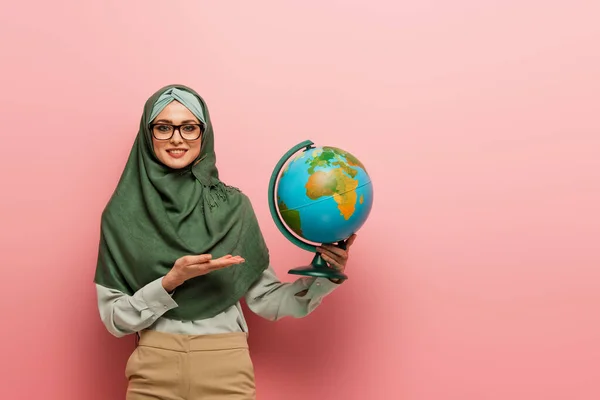 Professeur musulman souriant en hijab vert pointant vers le globe sur fond rose — Photo de stock