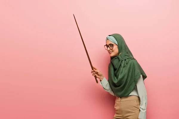 Sonriente maestro musulmán mirando hacia otro lado mientras sostiene el palo de apuntar sobre el fondo rosa - foto de stock