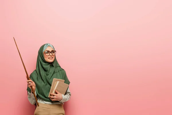 Joven profesor árabe en hijab verde sosteniendo palo de apuntar y libros sobre fondo rosa - foto de stock