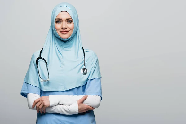 Alegre médico musulmán en hijab azul mirando a la cámara mientras está de pie con los brazos cruzados aislados en gris - foto de stock