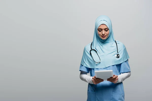 Médico árabe em hijab azul e uniforme olhando para comprimido digital isolado em cinza — Fotografia de Stock