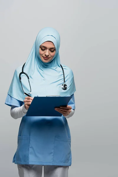 Medico abbastanza musulmano in hijab e scrittura uniforme sugli appunti isolati su grigio — Foto stock