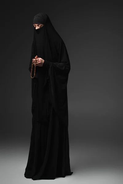 Full length view of muslim woman in black niqab praying with rosary on dark grey background — Stock Photo