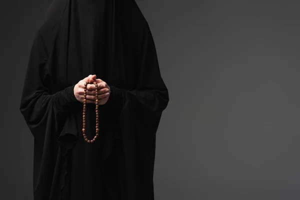 Частичный вид мусульманской монахини в черной Абайе, держащей молитвенные бусины изолированные на темно-серой — стоковое фото