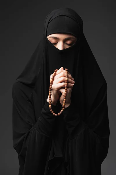 Jovem mulher muçulmana com olhos fechados segurando rosário enquanto reza isolado em preto — Fotografia de Stock
