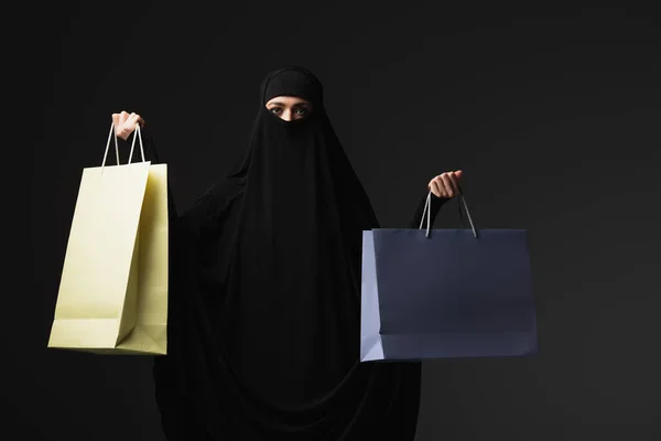 Muslim nun in black niqab holding multicolored shopping bags isolated on black — Stock Photo