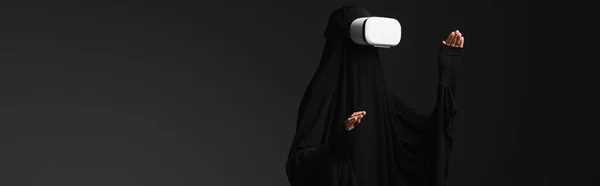 Muslim nun gesturing while gaming in vr headset isolated on black, banner — Stock Photo