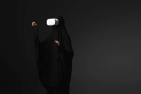 Monja musulmana en abaya negra haciendo gestos en auriculares vr aislados en negro - foto de stock