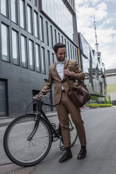 Longitud completa de hombre de negocios feliz en traje con bolsa de cuero y sonriendo cerca de la bicicleta fuera - foto de stock