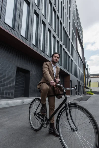 Full length of young businessman in suit with leather bag riding bicycle outside — Stock Photo