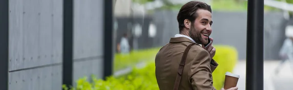 Felice giovane uomo d'affari in abbigliamento formale parlando sul cellulare e tenendo tazza di carta vicino all'edificio, banner — Foto stock