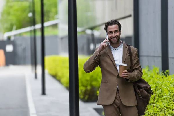 Cheerful businessman in formal wear talking on cellphone and holding paper cup near building — Stock Photo