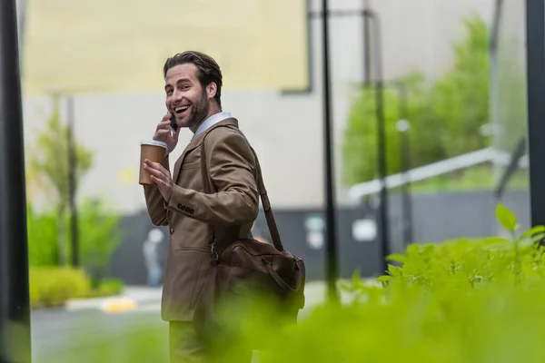 Cheerful businessman in formal wear talking on smartphone and holding paper cup outside — Stock Photo