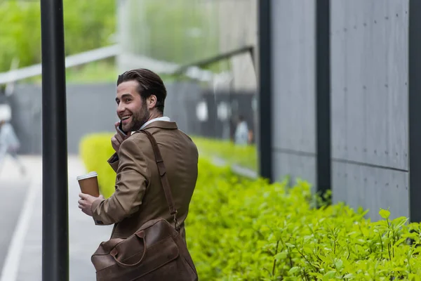 Homem de negócios sorridente em desgaste formal falando no celular e segurando copo de papel perto do edifício — Stock Photo