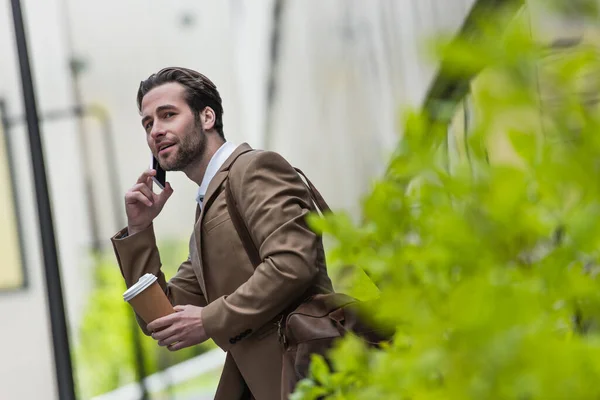 Улыбающийся мужчина в формальной одежде разговаривает по телефону и держит бумажную чашку рядом с размытым растением — стоковое фото