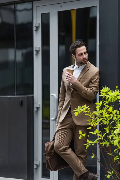 Pensive businessman in formal wear holding coffee to go near building — Stock Photo
