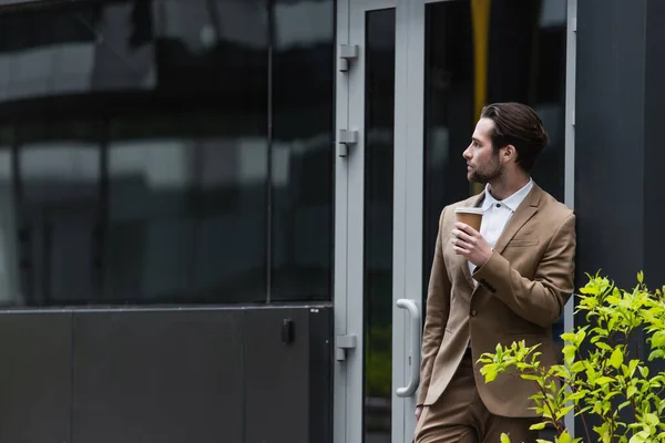 Thoughtful businessman in formal wear holding coffee to go near building — Stock Photo