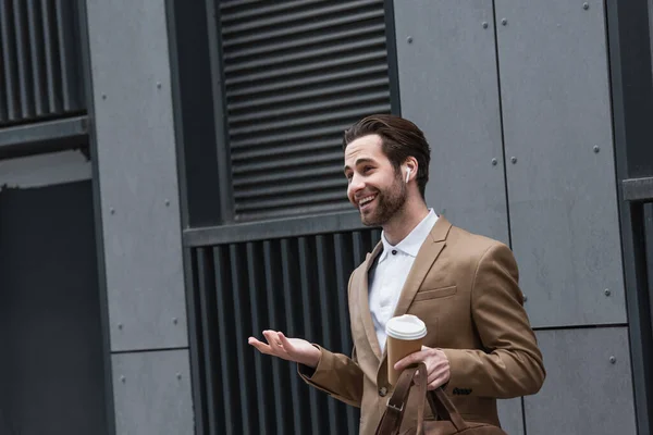 Feliz hombre de negocios en auriculares sosteniendo café para ir cerca de la construcción - foto de stock