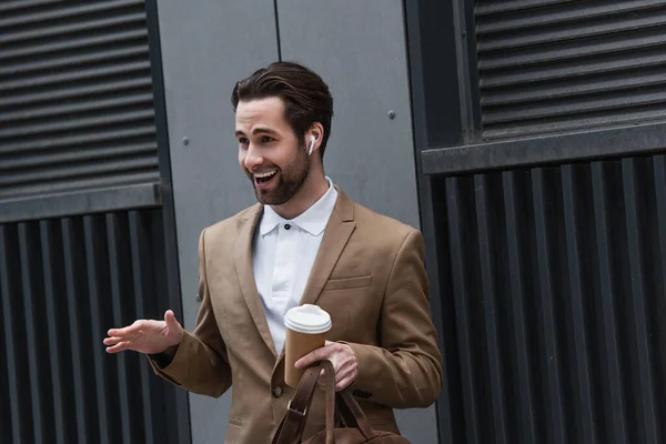 Cheerful businessman in earphones holding coffee to go near building — Stock Photo