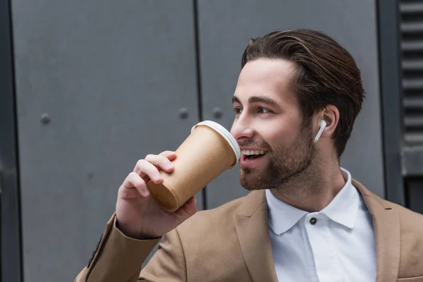 Happy businessman in earphones drinking coffee to go near building — Stock Photo