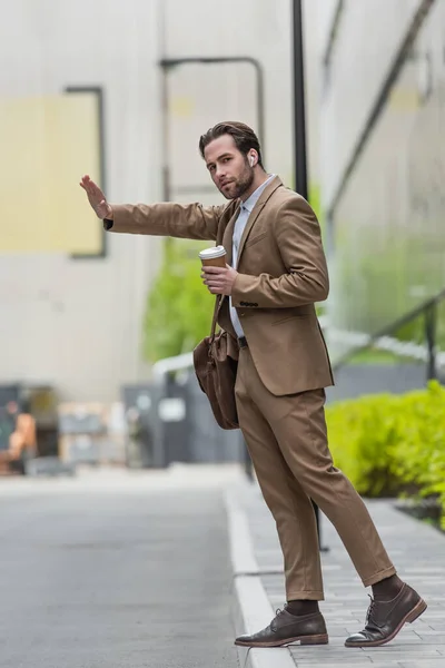 Full length of businessman in earphones holding coffee to go and gesturing while catching cab — Stock Photo