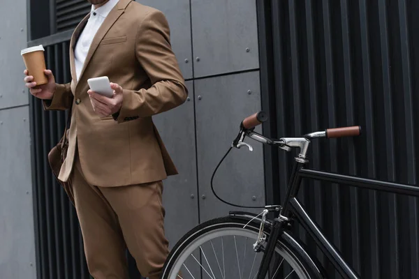 Cropped view of businessman holding paper cup and smartphone near bicycle — Stock Photo
