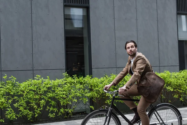 Jovem empresário em fones de ouvido andar de bicicleta perto do edifício — Fotografia de Stock