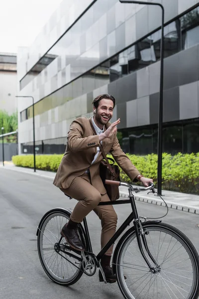 Full length of cheerful businessman in earphones riding bicycle and waving hand near building and plants — Stock Photo