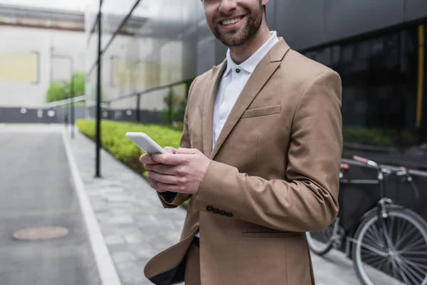 Cropped view of bearded businessman smiling while holding smartphone outside — Stock Photo