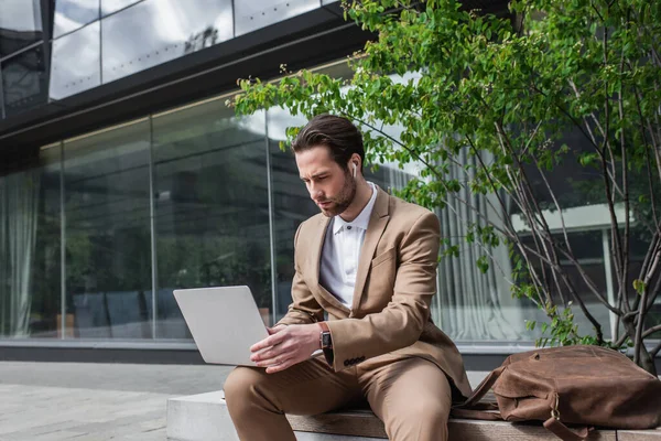 Businessman in earphones using laptop while sitting on bench — Stock Photo