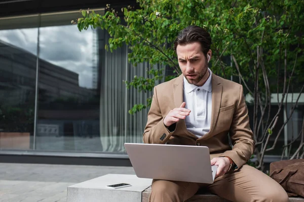 Hombre de negocios en auriculares usando el ordenador portátil mientras gesticulaba y se sentaba en el banco - foto de stock