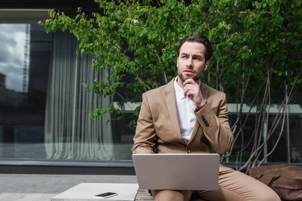 Homem de negócios pensativo em fones de ouvido usando laptop enquanto sentado no banco — Fotografia de Stock