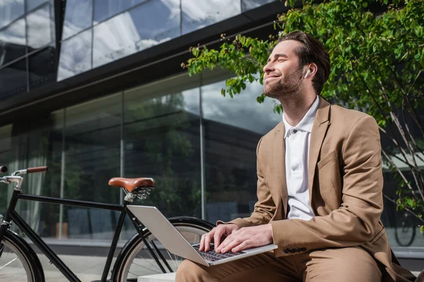 Pleased businessman in earphones using laptop while sitting on bench — Stock Photo