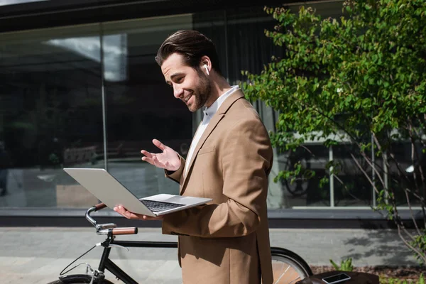 Happy businessman in earphones having video call on laptop outside — Stock Photo
