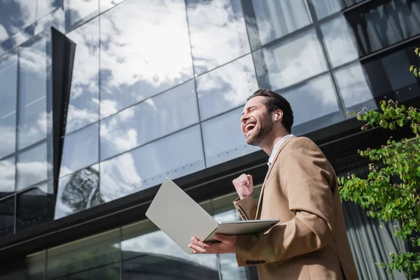 Vue à angle bas de l'homme d'affaires joyeux dans les écouteurs tenant ordinateur portable à l'extérieur — Photo de stock