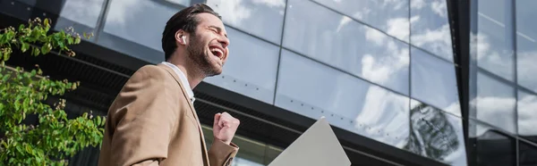 Blick auf einen fröhlichen Geschäftsmann mit Kopfhörer und Laptop draußen, Banner — Stockfoto