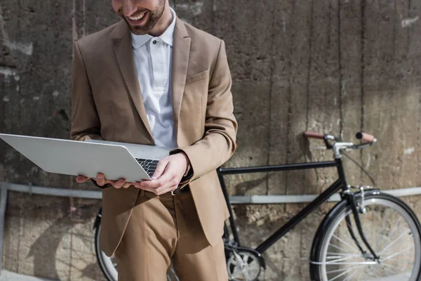 Partial view of cheerful businessman holding laptop near building and bicycle outside — Stock Photo