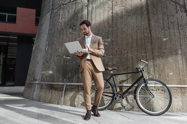 Volle Länge des Geschäftsmannes in Kopfhörer halten Laptop in der Nähe Gebäude und Fahrrad außerhalb — Stockfoto