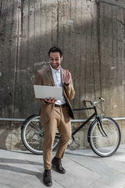 Full length of happy businessman in earphones holding laptop while having video call outside — Stock Photo